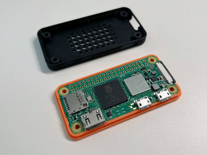 A Raspberry Pi board in an orange protective case, shown with its black plastic back cover placed above it on a light gray surface.