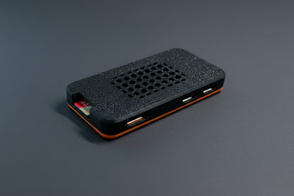 A small rectangular electronic device with a textured black top, a grid of circular cutouts, and visible USB-A ports on the side, photographed on a plain light background.