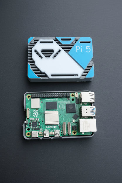 A Raspberry Pi 5 board inside a small gray protective case, shown below its blue and white branded protective cover on a textured concrete surface.