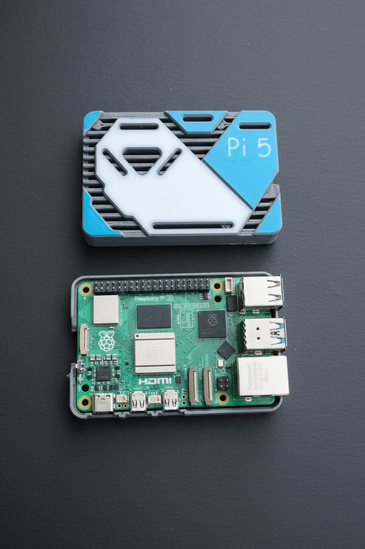 A Raspberry Pi 5 board inside a small gray protective case, shown below its blue and white branded protective cover on a textured concrete surface.